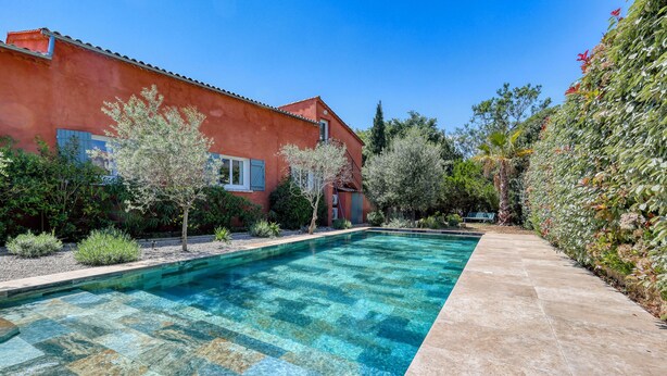 A swimming pool with clear blue water, surrounded by a red building with a tiled roof, and a garden with various plants and trees. - Large villa for 2 families - 6 bedrooms - Secure swimming pool - Six-Fours (Six fours les plages)