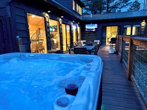 A hot tub with a glass and a cup, a wooden deck, and a building with a TV. - New Luxury Cabin/Hot Tub/Custom Playset/Firepit/Hammocks/Corn Hole/Games (Broken Bow)