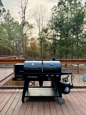 A barbecue grill on a wooden deck with a view of trees and a fence in the background. - New Luxury Cabin/Hot Tub/Custom Playset/Firepit/Hammocks/Corn Hole/Games (Broken Bow)