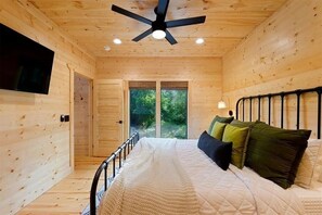 A wooden cabin bedroom with a bed, a ceiling fan, a TV, and a window with a view of greenery. - New Luxury Cabin/Hot Tub/Custom Playset/Firepit/Hammocks/Corn Hole/Games (Broken Bow)