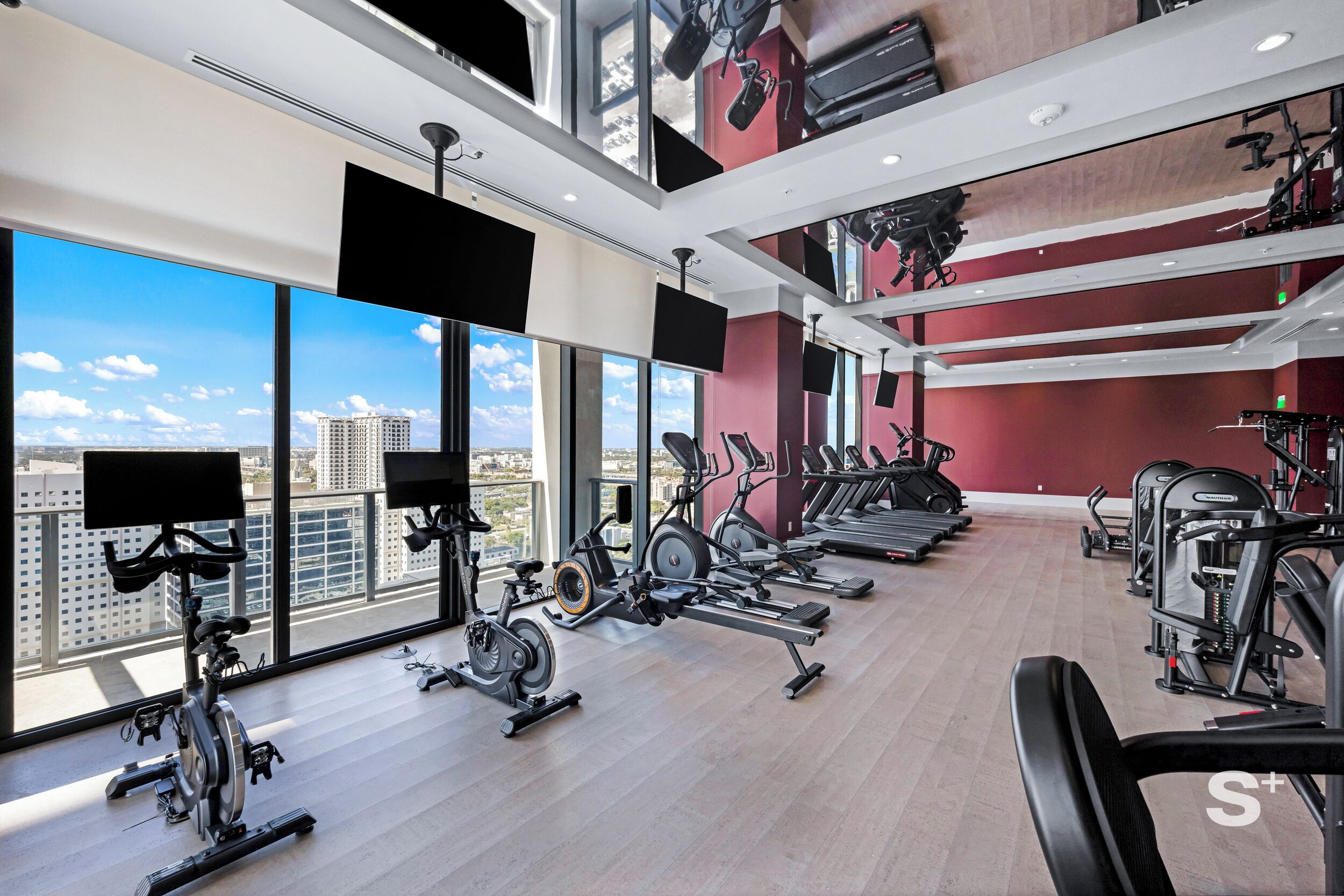 A modern gym with various exercise equipment, large windows offering city views, and a high ceiling.