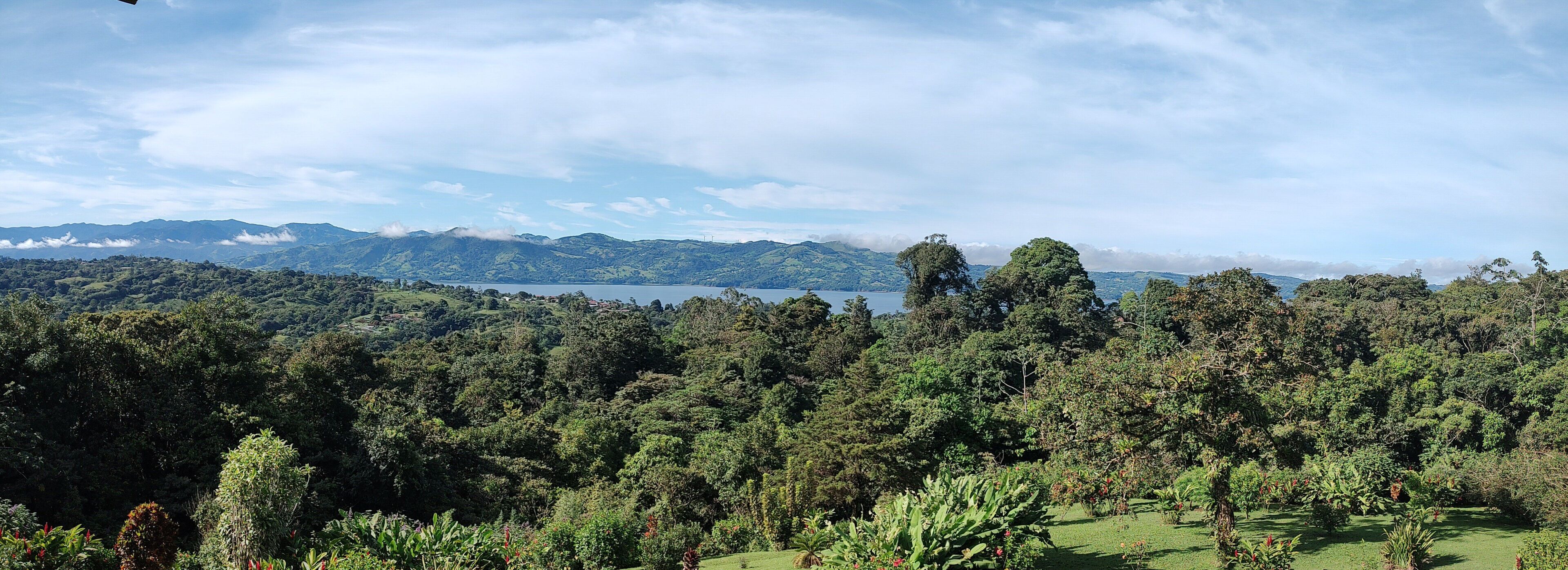 A lush landscape with dense greenery, a lake, and distant mountains under a partly cloudy sky.