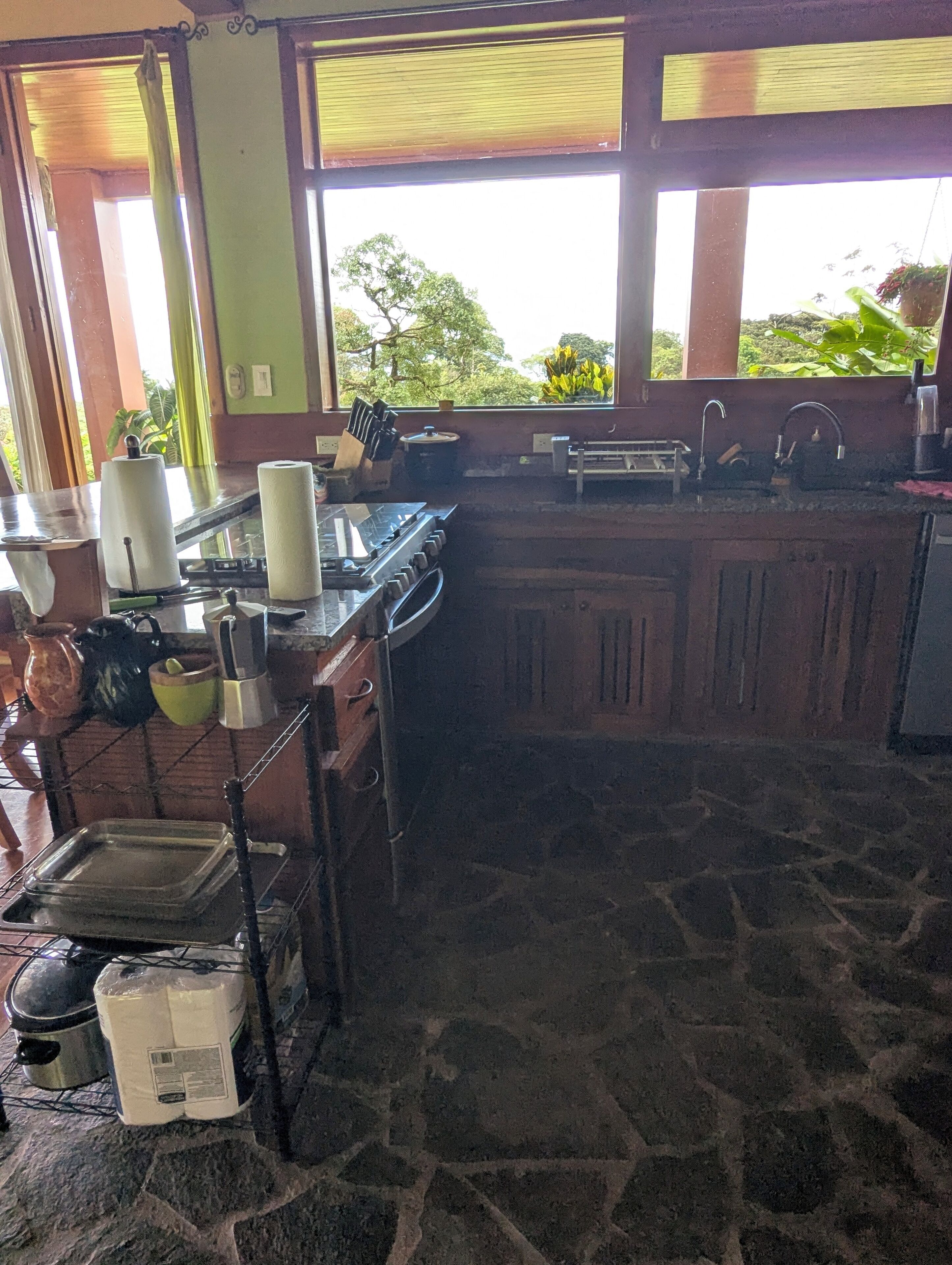 A kitchen with a stone floor, a stove, a sink, and a window with a view of greenery.