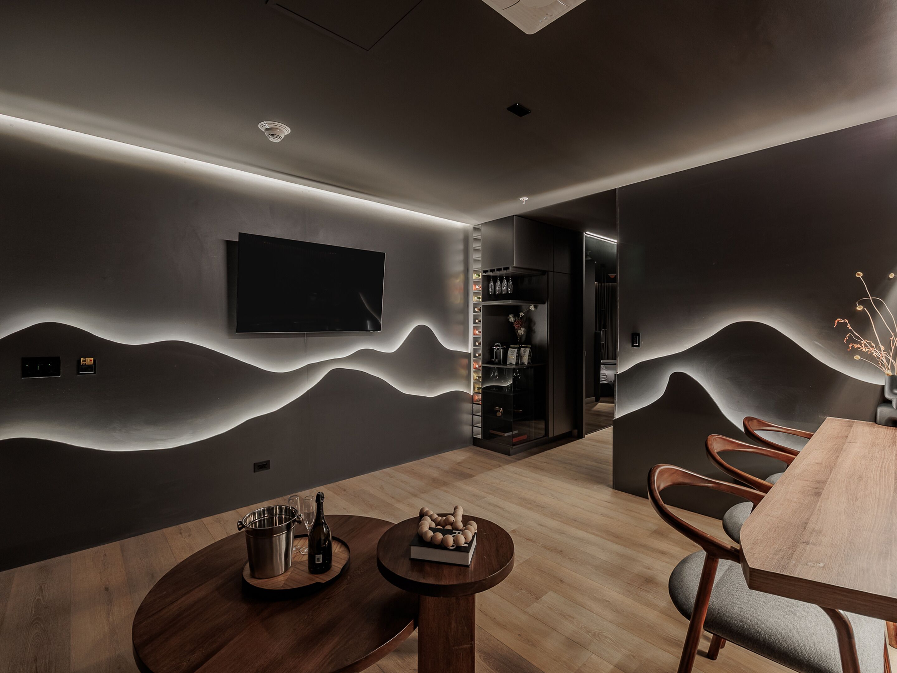 A modern dining area with a wooden table and chairs, a mounted flat-screen TV, and a wall-mounted shelf with bottles.