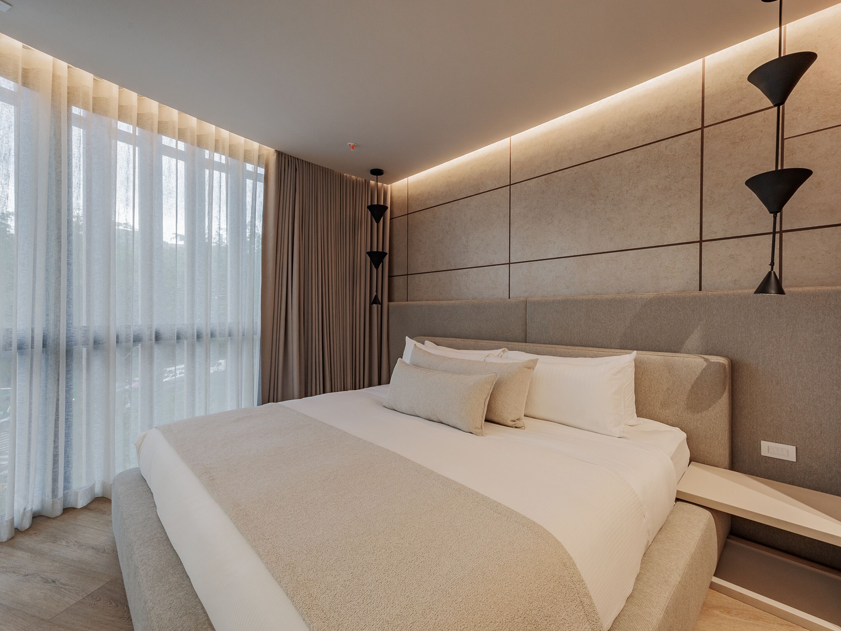 A modern bedroom with a large bed, a bedside table, a wall-mounted lamp, and a view through sheer curtains.