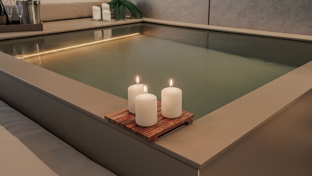 Ett modernt spa med ett rektangulärt bubbelbad, tre tända ljus på ett träbricka och en beige soffa i bakgrunden.
