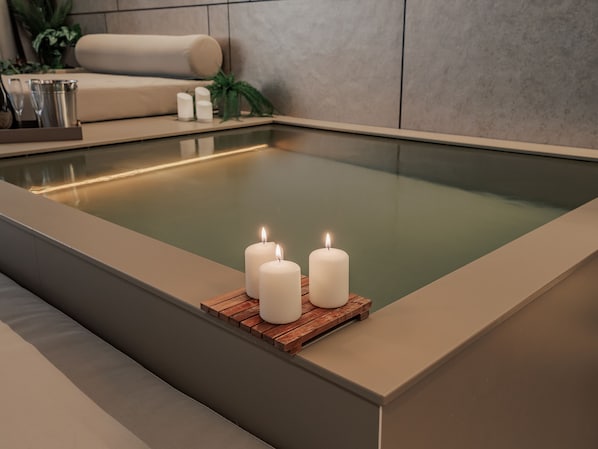 A modern spa with a rectangular hot tub, three lit candles on a wooden tray, and a beige sofa in the background. - Devú (Medellín)