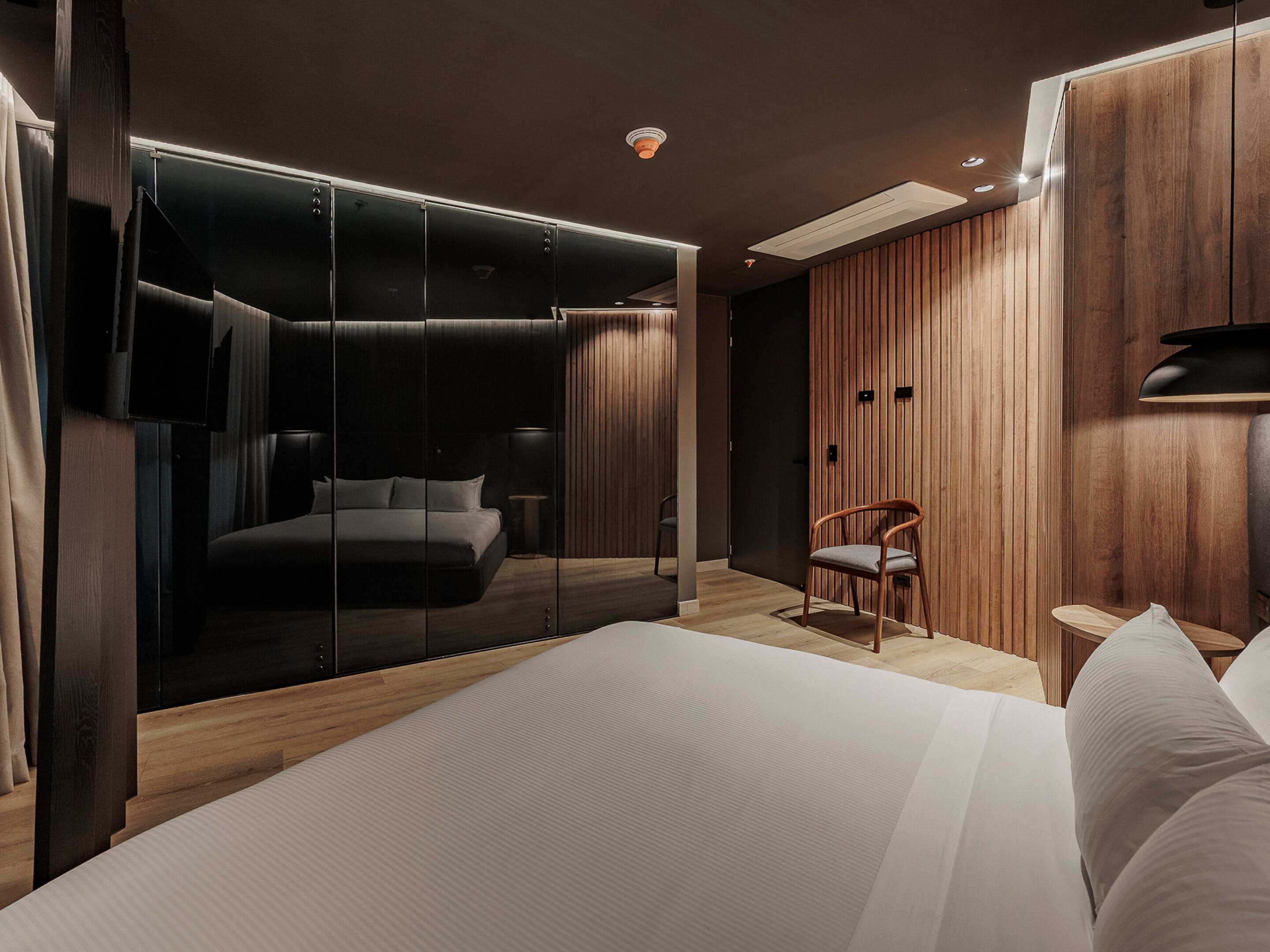 A modern bedroom with a large bed, a wooden wardrobe, a chair, and a TV mounted on the wall.