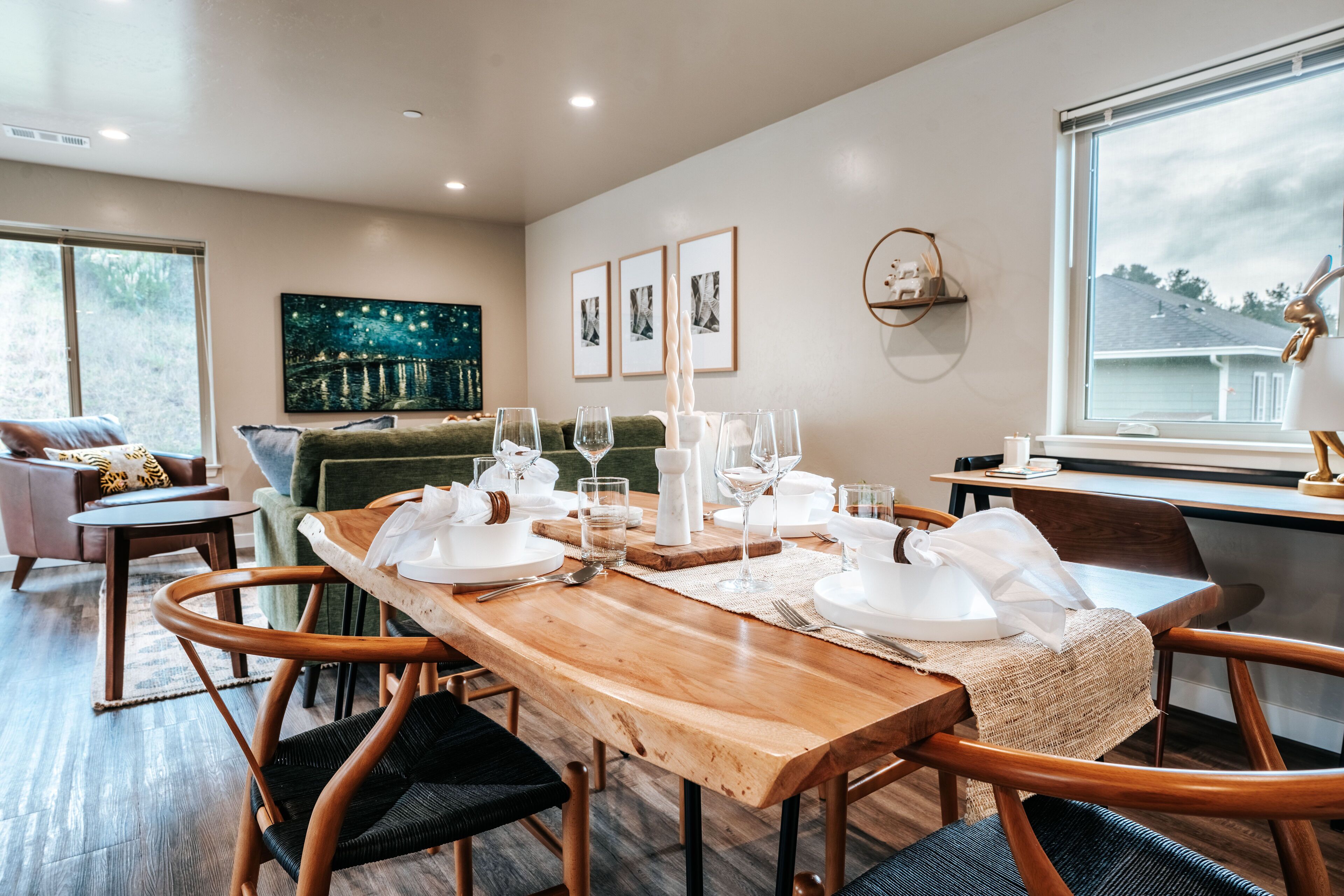 A dining area with a wooden table set for four, surrounded by wicker chairs. There are framed pictures on the wall, a window with a view of the outside, and a green sofa in the background.
