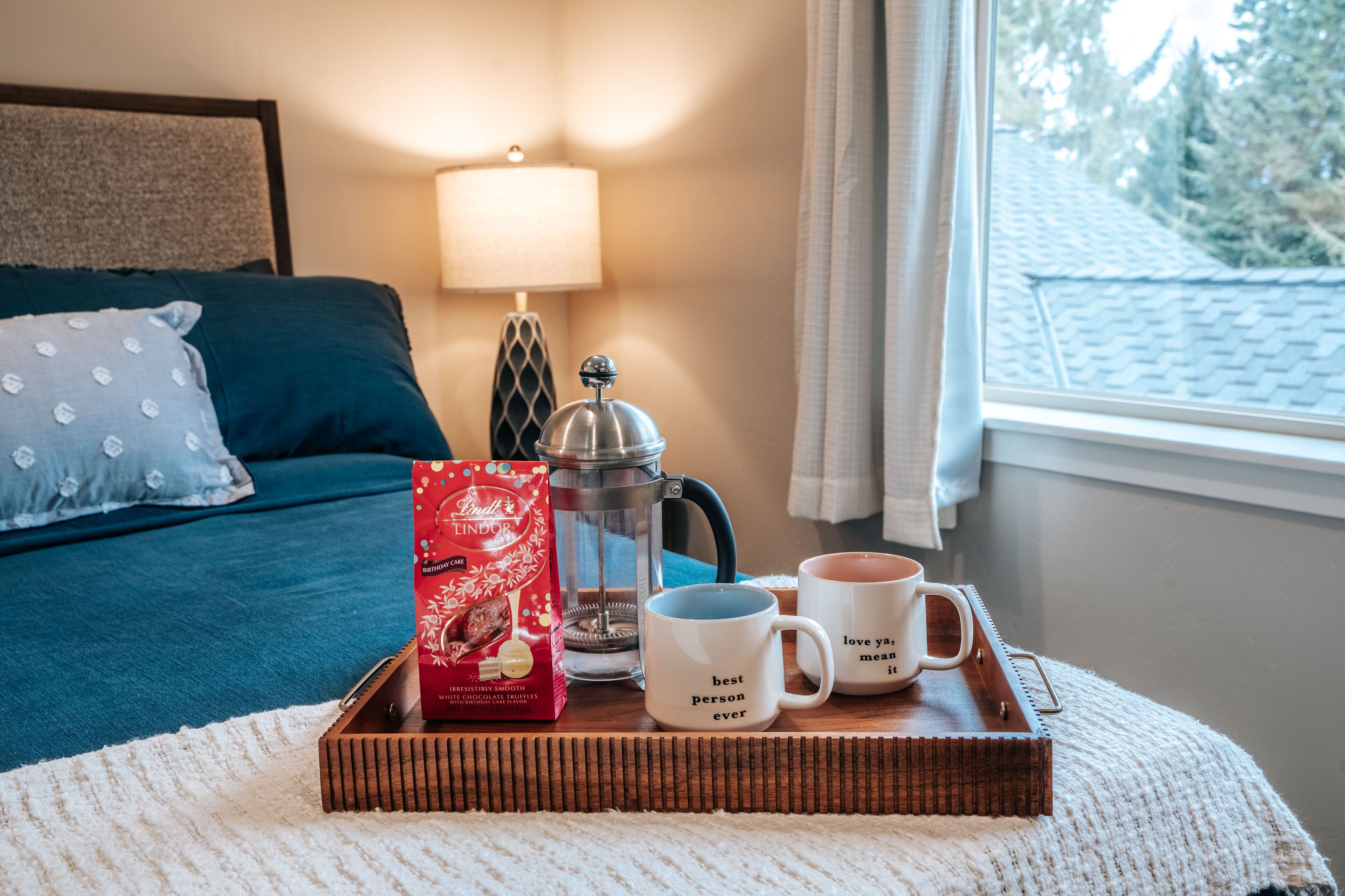 A cozy hotel room with a bed, a bedside table with a lamp, a tray with a coffee pot, two mugs, and a box of Lindt chocolates.