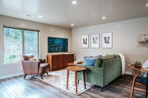 A modern living room with a leather armchair, a green sofa, a wooden table, and a large window with a view of greenery.