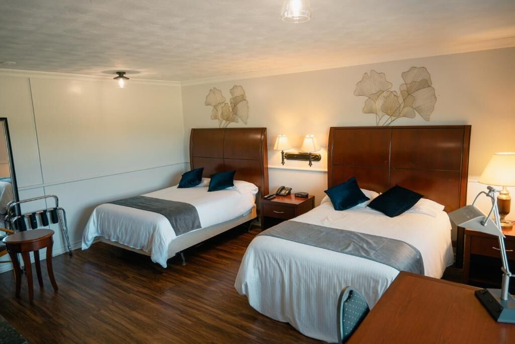 A hotel room with two beds, wooden headboards, bedside tables with lamps, a small round table, and a chair.