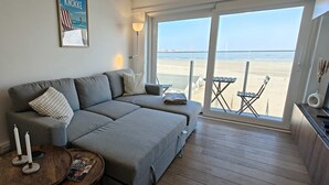 A living room with a grey sofa, a small round table with candles, a magazine, and a view of the beach through large windows. - Zeezicht studio met aparte slaaphoek (Knokke-Heist)