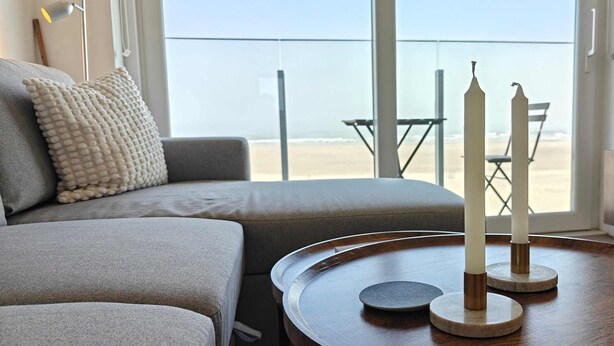 A modern living room with a sofa, a round wooden table with candles, and a view of the beach through large windows. - Zeezicht studio met aparte slaaphoek (Knokke-Heist)