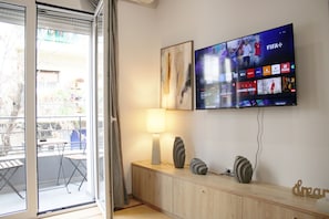 Una habitación con una televisión mostrando un canal deportivo, un aparador de madera con adornos, una mesa con sillas y una puerta corredera de vidrio que da a un espacio exterior.