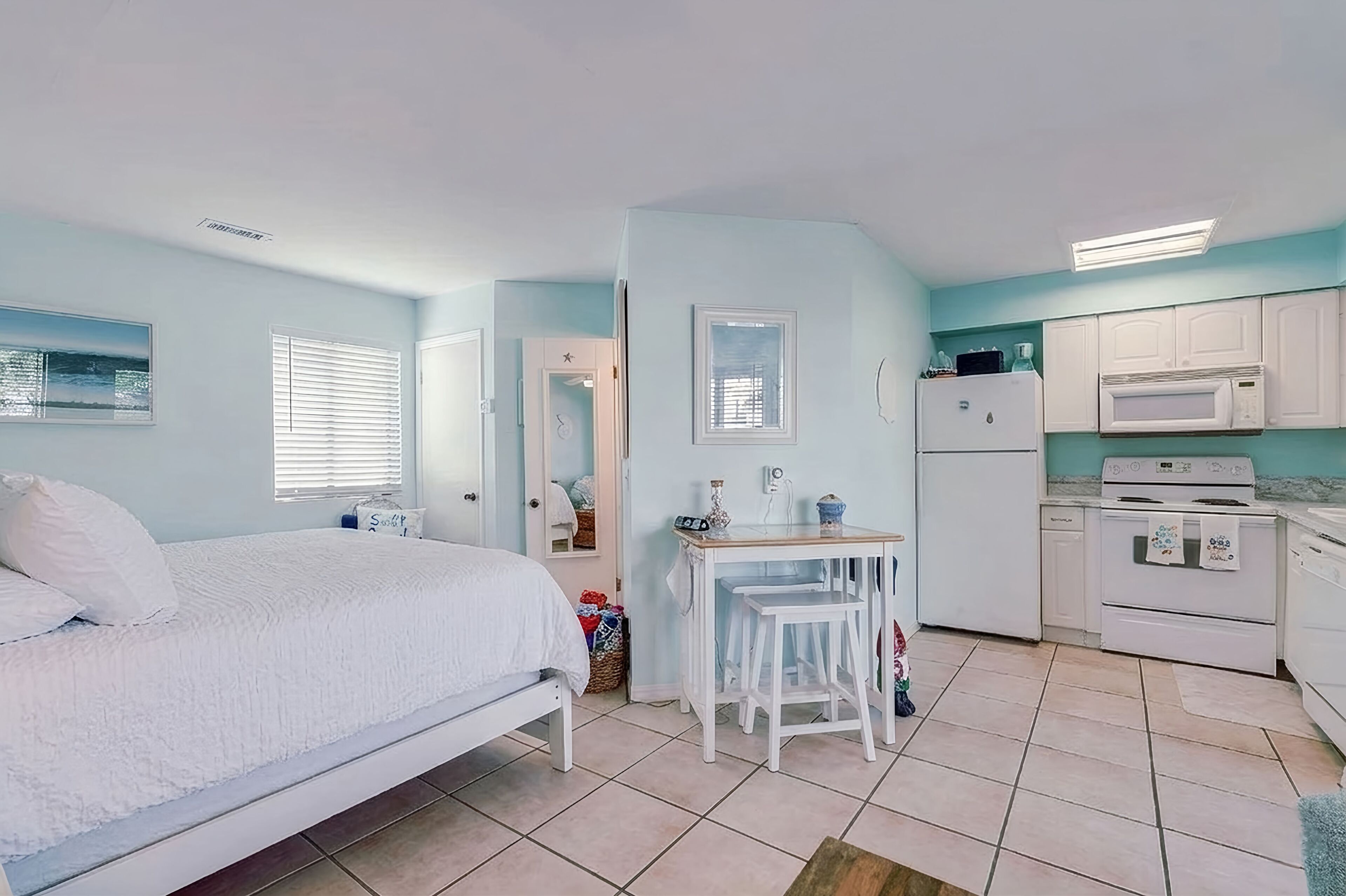 Un dormitorio con cama, una mesita con sillas, una ventana con persianas y una zona de cocina con refrigerador, microondas y horno.