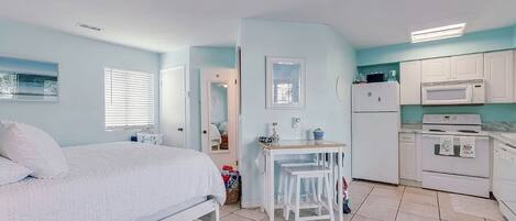 Un dormitorio con cama, una mesita con sillas, una ventana con persianas y una zona de cocina con refrigerador, microondas y horno.