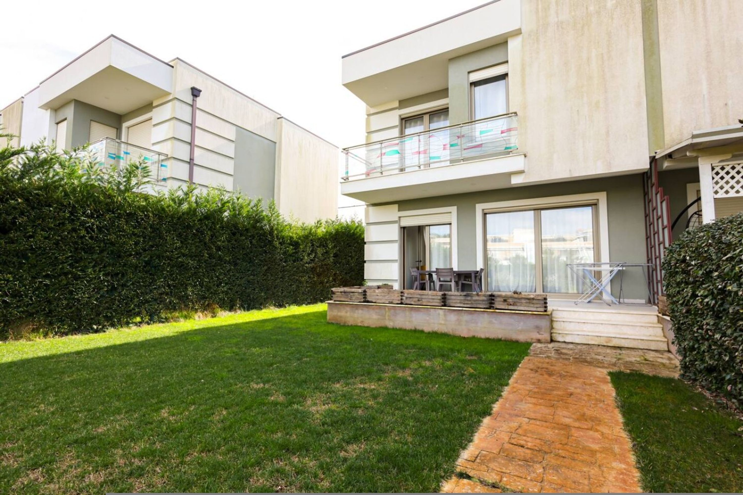 A modern two-story residential building with a flat roof, large windows, and a balcony with a glass railing.