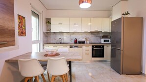 Una cocina moderna con armarios blancos, un refrigerador de acero inoxidable y una encimera de mármol. Hay una mesa de comedor con sillas y una lámpara colgante arriba.