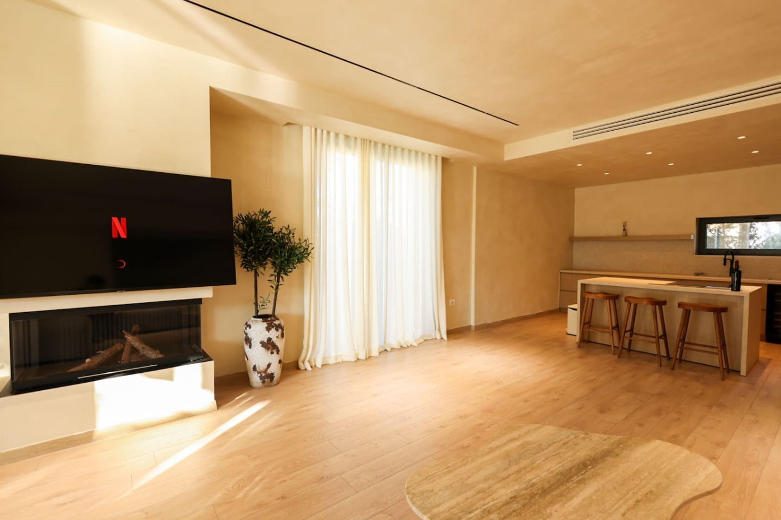 A modern living room with a flat-screen TV, a fireplace, a dining area with bar stools, and a large window with curtains.
