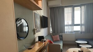 Quarto de hotel com escrivaninha de madeira, espelho redondo, sofá, cama com toalhas, mesinha e janela com cortinas.