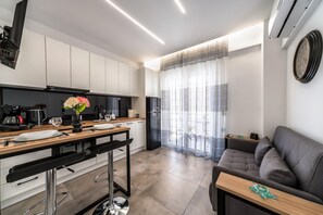 Une cuisine moderne avec des meubles blancs, un coin repas doté d’une table en bois et de chaises noires, et un canapé gris dans un salon offrant une vue à travers des rideaux transparents.