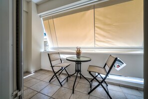 A balcony with a table and two chairs, a window with a beige blind, and a small plant on the table. - Nicol studio by Cretanhouses (Iraklio)