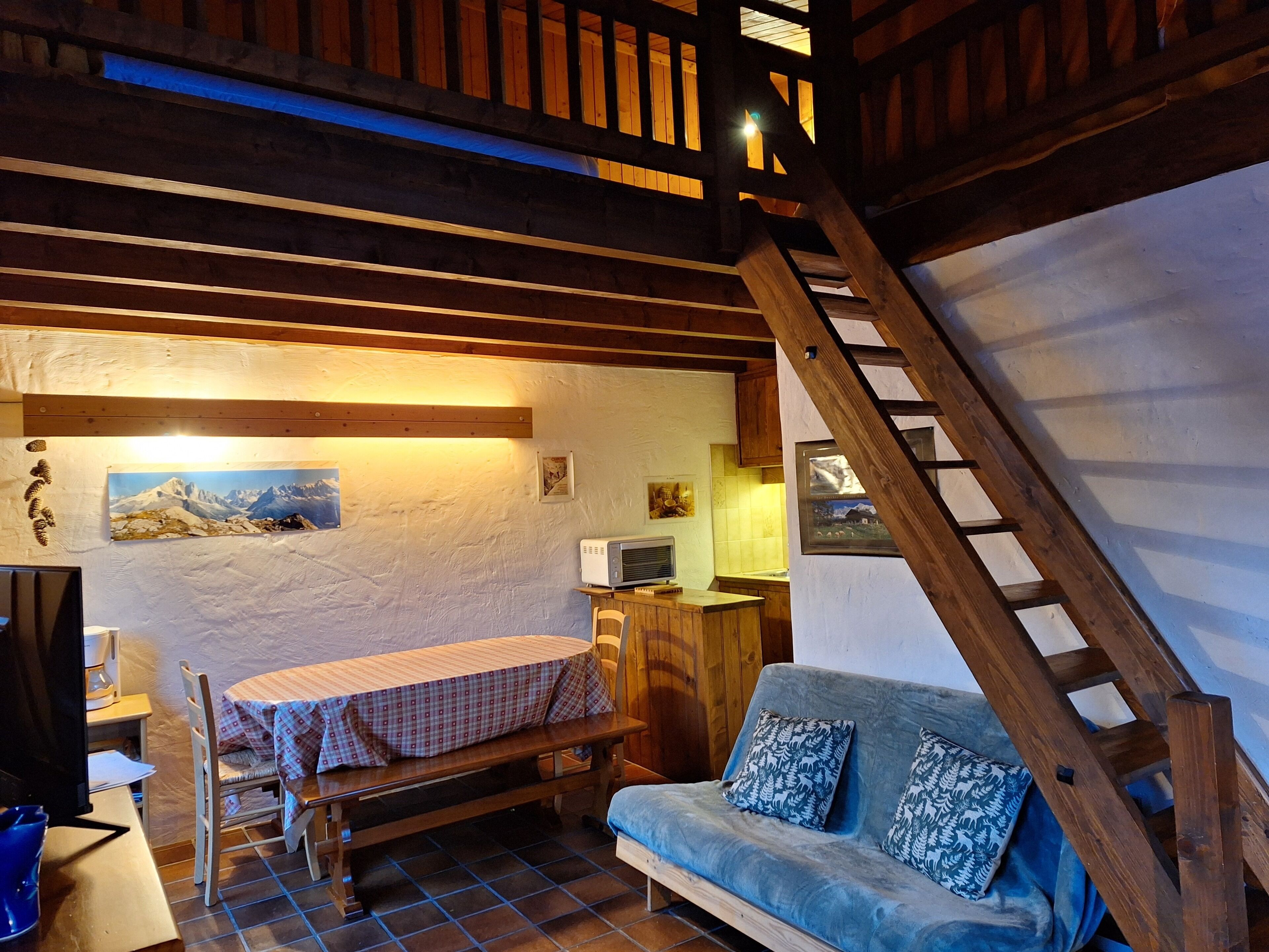 Ein gemütlicher Raum mit einer Holztreppe, einem blauen Sofa, einem kleinen Tisch mit kariertem Tischläufer und einem an der Wand befestigten Fernseher.
