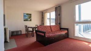 A living room with a sofa, a dining table, chairs, a painting, and large windows. - Charming apartment for 4 people near shops (Grasse)