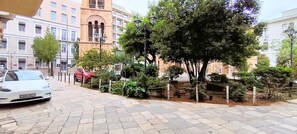 Eine gepflasterte Straße mit einem weißen Auto, das links parkt, einem roten Auto im Hintergrund und einem großen Baum im Vordergrund.