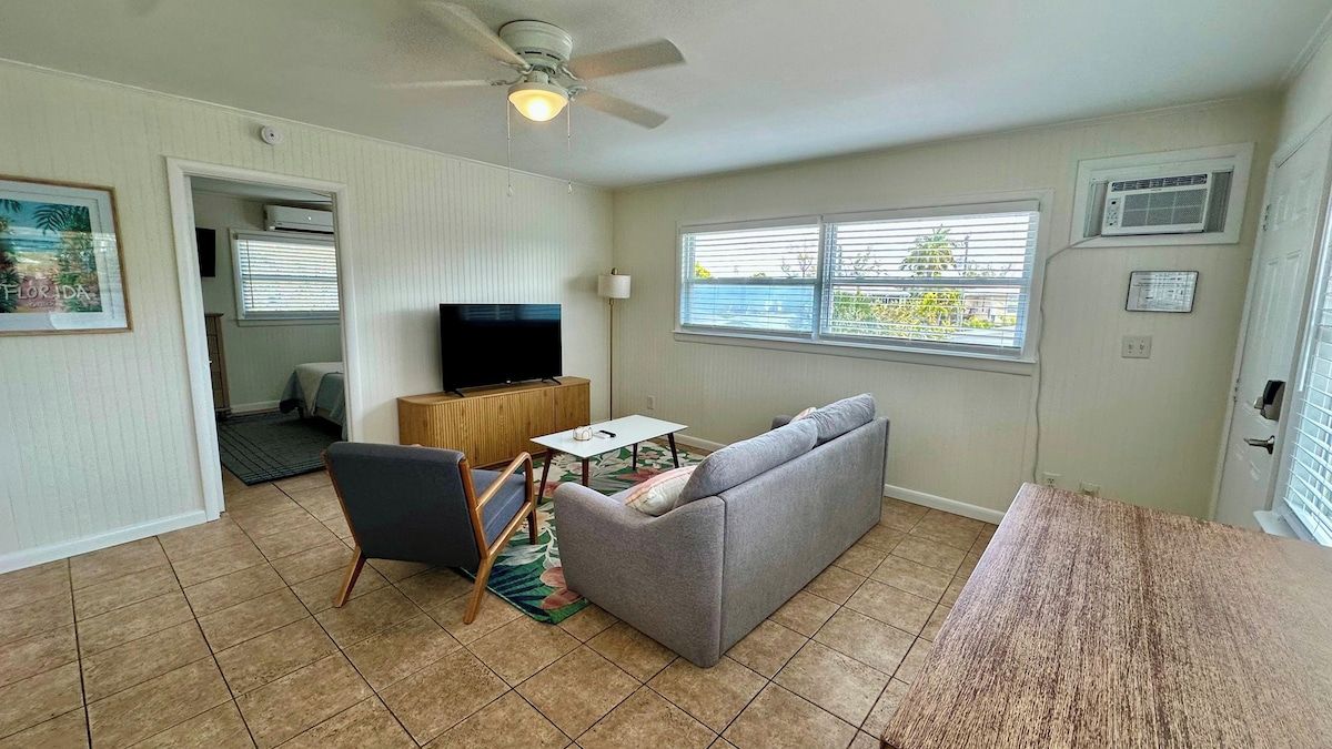A living room with a sofa, a television, a ceiling fan, and a window with blinds.