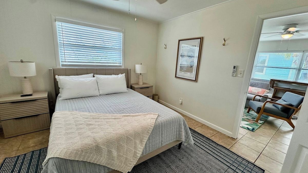 A bedroom with a bed, bedside tables, a window with blinds, a framed picture on the wall, and a chair in the adjacent room.