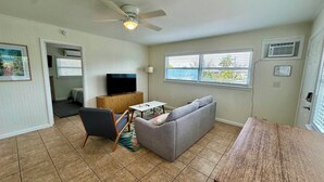 Ein Wohnzimmer mit grauem Sofa, einem Holztisch, einem schwarzen Sessel, einem Deckenventilator und einem Fernseher auf einem Holzschrank.