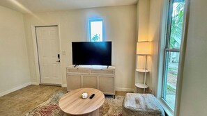 Ein Wohnzimmer mit Flachbildfernseher auf einer Kommode, einem runden Couchtisch, einer Lampe und einem Fenster mit Blick ins Grüne.