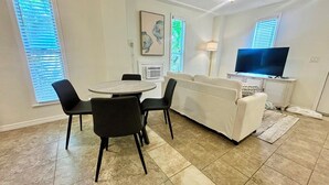 Ein Wohnzimmer mit einem weißen Sofa, einem runden Esstisch mit Stühlen, einem Flachbildfernseher und einem Gemälde an der Wand.