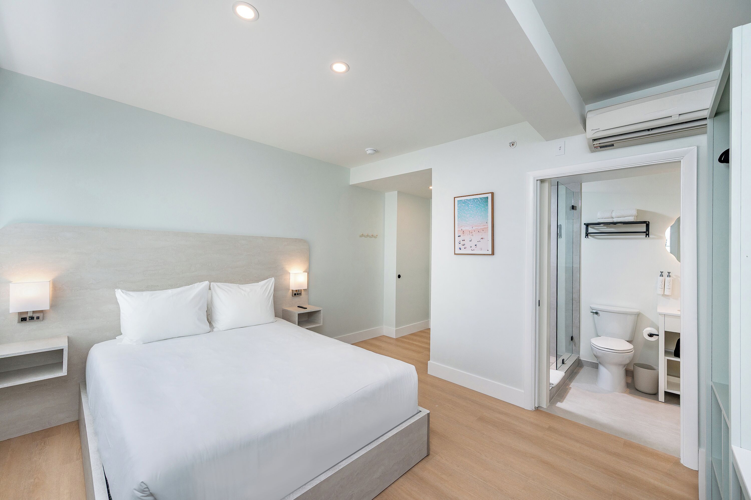 A modern hotel room with a large bed, bedside lamps, a bathroom with a toilet and sink, and an air conditioning unit.