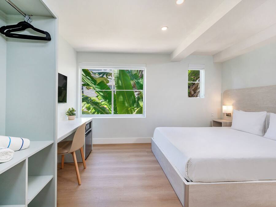 A bedroom with a bed, desk, chair, and a window with a view of greenery.