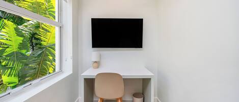 A corner room with a white desk, a chair, a TV mounted on the wall, a window with a view of green foliage, and a trash bin.
