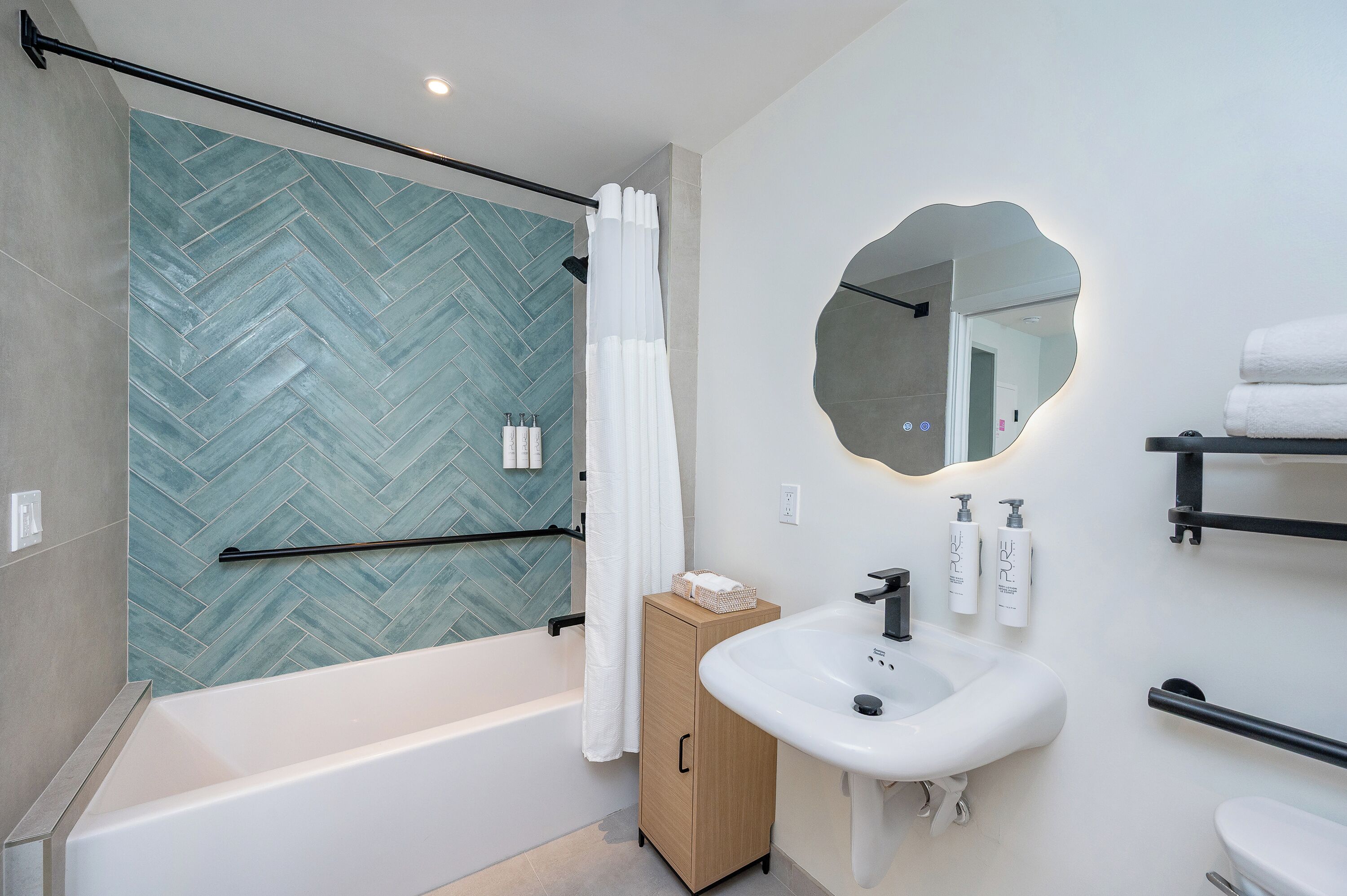 Un baño moderno con pared de ducha con azulejos de espiga, una bañera independiente, un mueble de madera, un lavabo blanco y un espejo redondo.