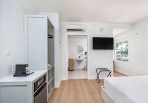 A modern hotel room with a kitchenette, a TV mounted on the wall, a window with a view, and a bathroom visible through an open door.