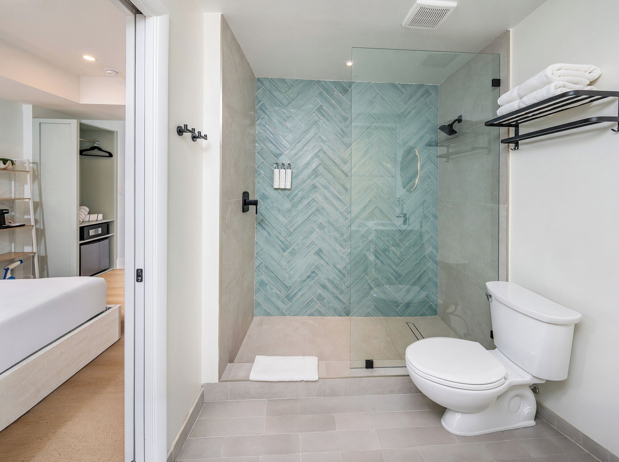 A modern bathroom with a glass shower enclosure, a toilet, and a bed in the adjacent room.