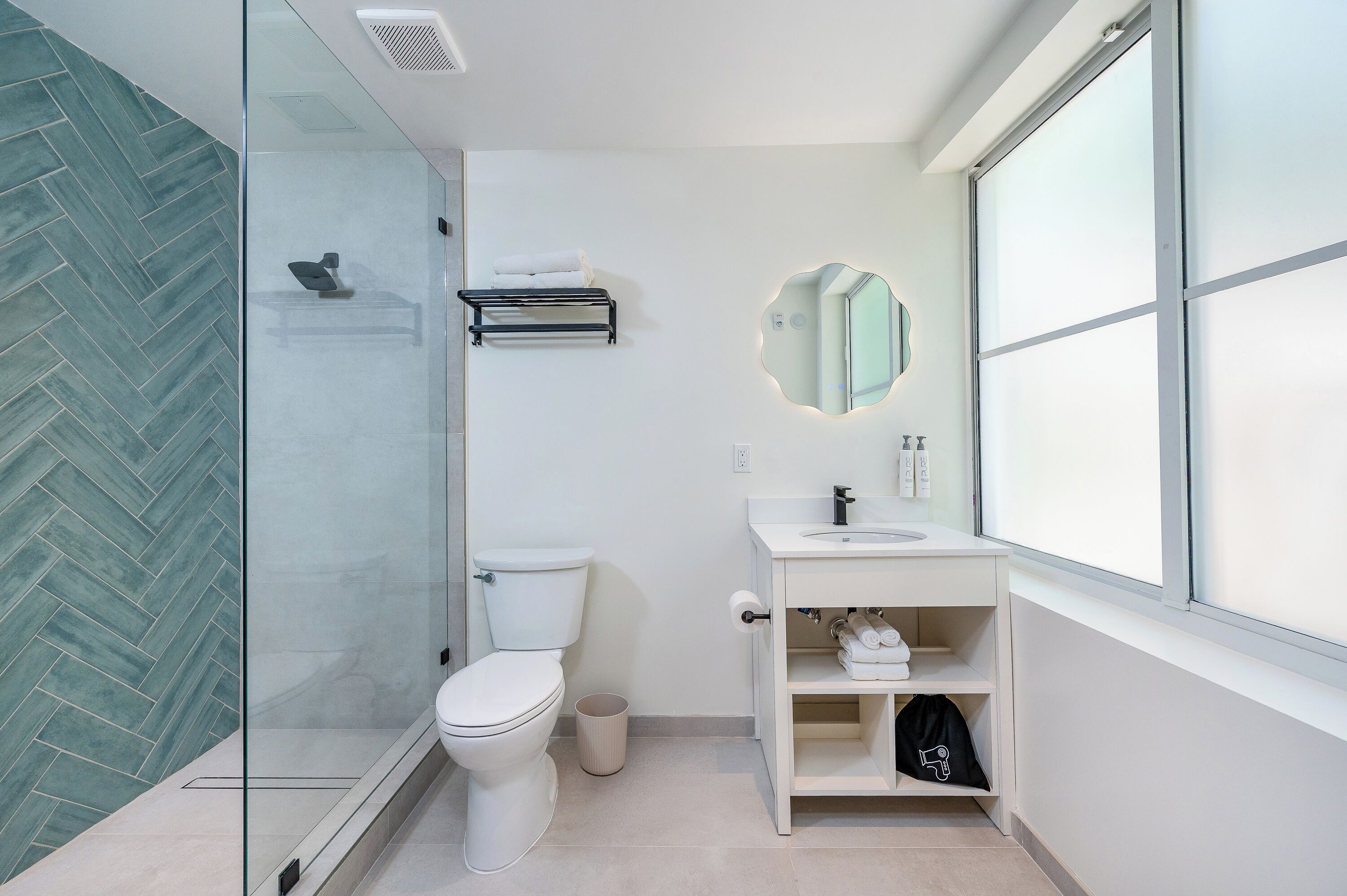 Et moderne badeværelse med glasvæg ved brusebad, toilet, vask med spejl og et vindue.