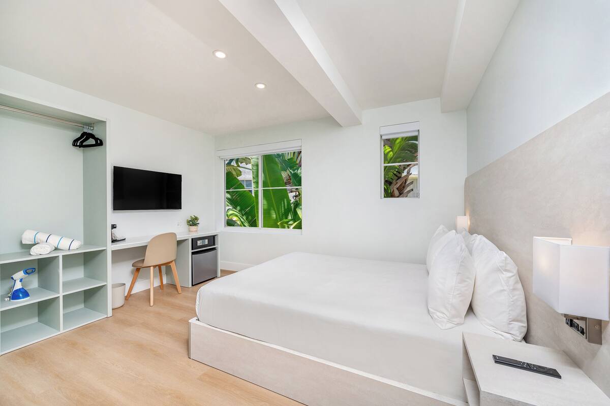 Un dormitorio moderno con una cama, un escritorio, una silla, un televisor y una ventana con vistas a la vegetación.
