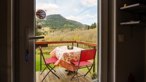 Un balcon avec une table et des chaises, une vue sur une colline boisée et une porte dotée d’une poignée en métal.