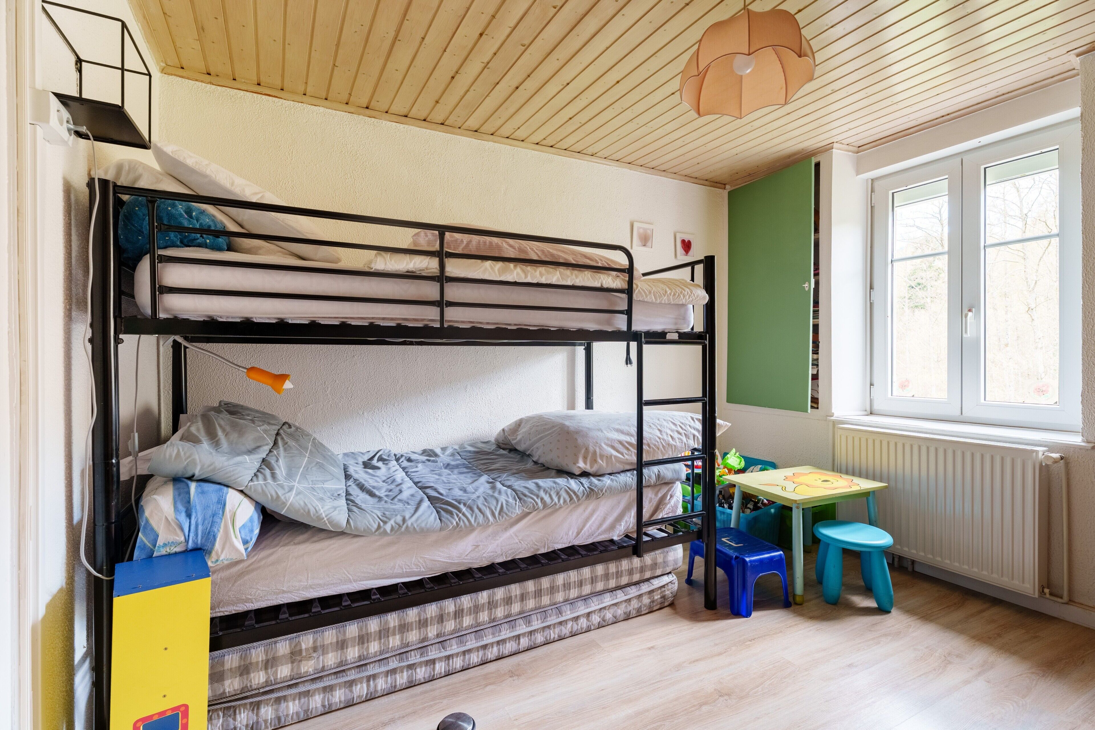 Een slaapkamer met een stapelbed, een tafel, krukjes, een radiator en een raam.
