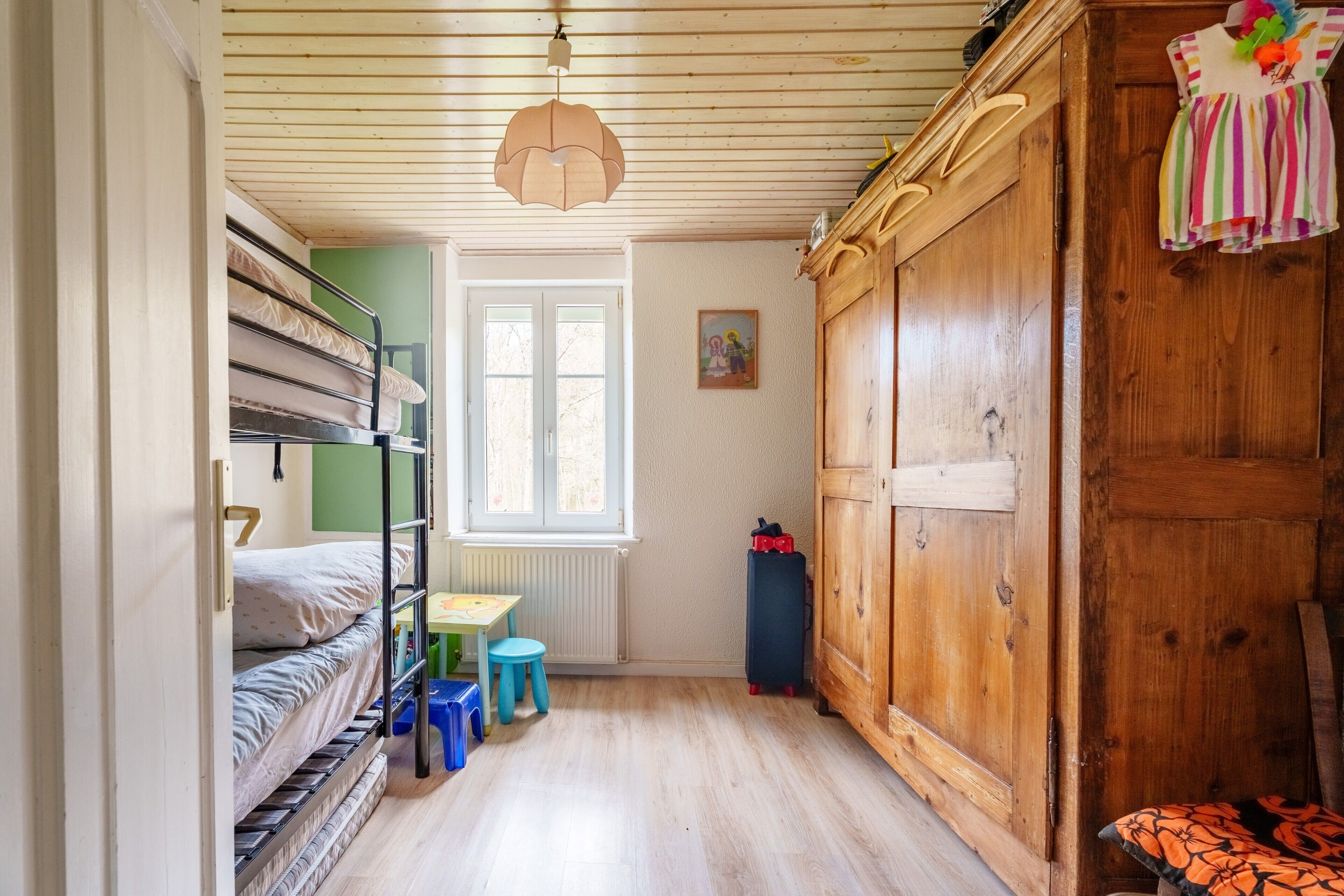 Une chambre avec des lits superposés, une armoire en bois, une petite table avec un tabouret et une fenêtre.