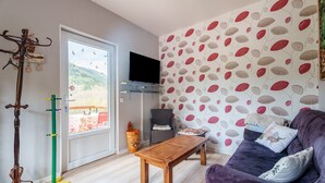 Un salon avec une table basse en bois, un canapé violet, un téléviseur posé sur un meuble, un porte-manteau et une fenêtre donnant sur un paysage verdoyant.