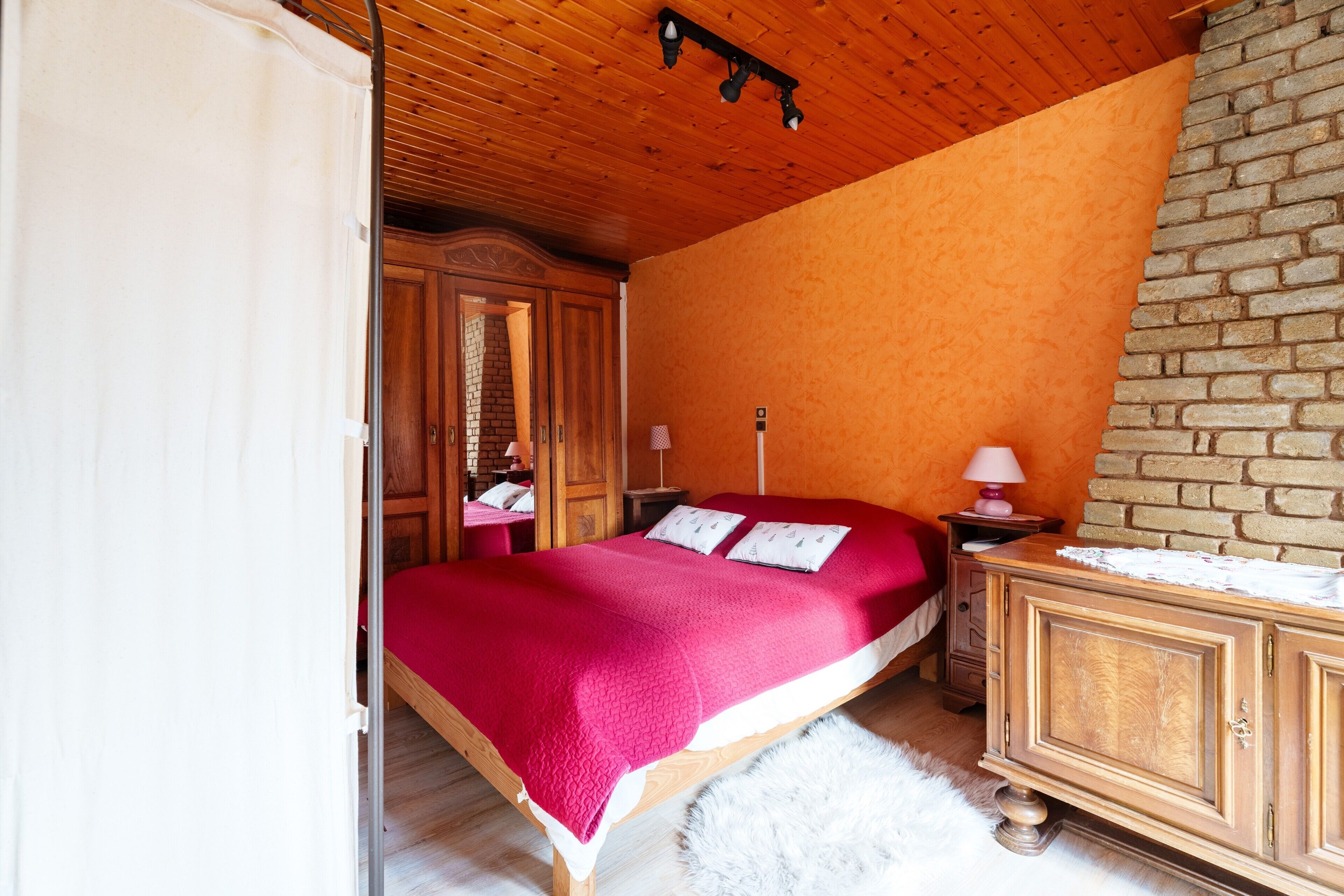 Une chambre avec un plafond en bois, un mur en briques, un lit recouvert d’une couverture rouge, une commode en bois et un tapis blanc et moelleux.