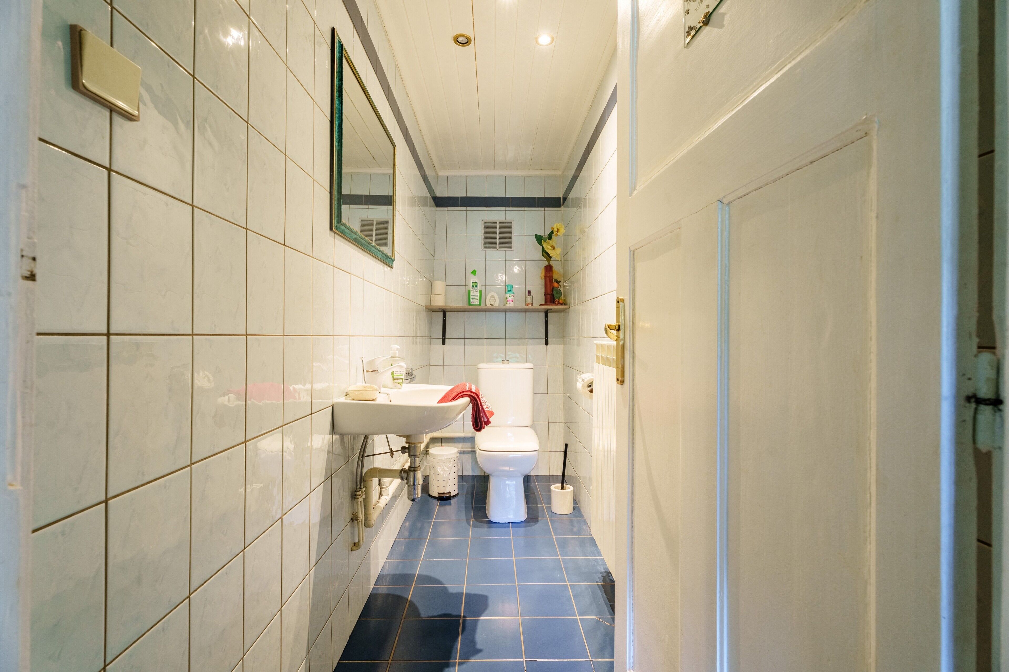 Une salle de bain avec des murs recouverts de carreaux blancs, un sol bleu, un lavabo blanc, des toilettes et un miroir.