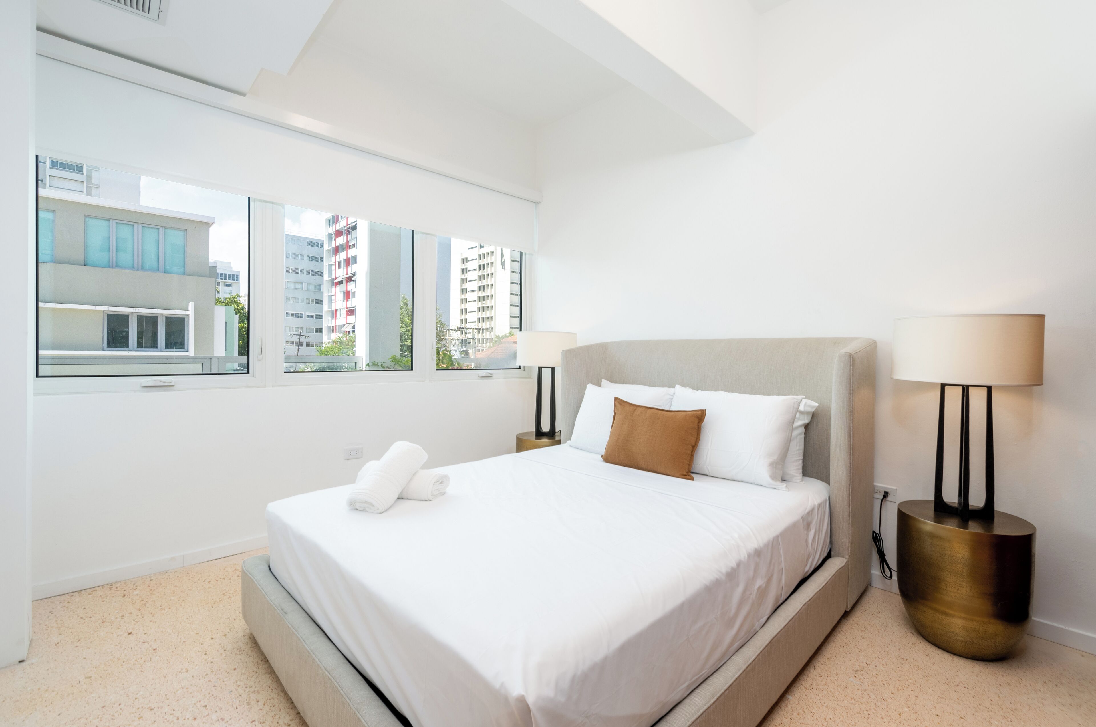 Habitación de hotel moderna con una cama grande, dos lámparas de noche, vistas de edificios por la ventana y una pared blanca.